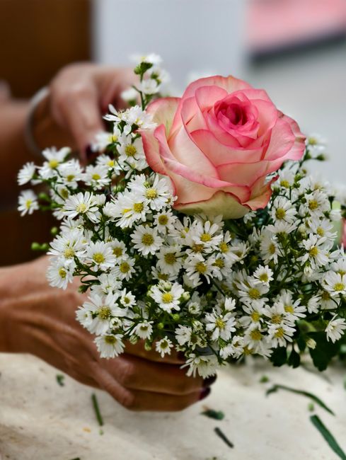 A person holding a small floral arrangement consisting of a single large pink and cream rose surrounded by a cluster of small white daisies with yellow centers. The bouquet is loosely assembled, with