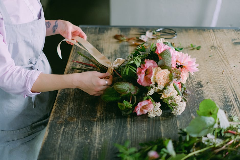 Close-up of a florist’s hands tying a beige ribbon around a freshly arranged bouquet of flowers on a rustic wooden table. The bouquet includes pink peonies, cream-colored roses, white waxflowers, an