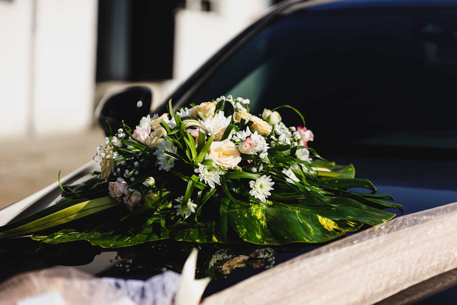A floral arrangement featuring a bouquet of white and pale pink roses, white daisies, and baby's breath, accented with lush green foliage. The bouquet is placed on the hood of a sleek black car, parti