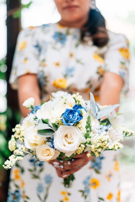 A woman holding a floral bouquet featuring white roses, blue roses, and small white and yellow accent flowers, with green and silver foliage, wrapped in white paper and tied with a white ribbon. The b