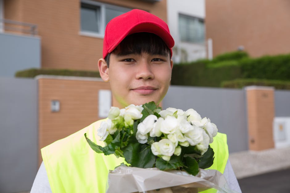 A young delivery person wearing a red cap and a neon yellow jacket holds a bouquet of fresh white roses wrapped in white paper, standing outdoors in front of modern residential buildings with brick an
