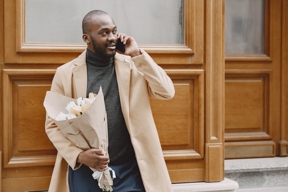 A man with dark skin, a beard, and a shaved head is standing outdoors in front of a wooden-paneled building, holding a bouquet of white flowers wrapped in brown paper with a white ribbon. He is wearin