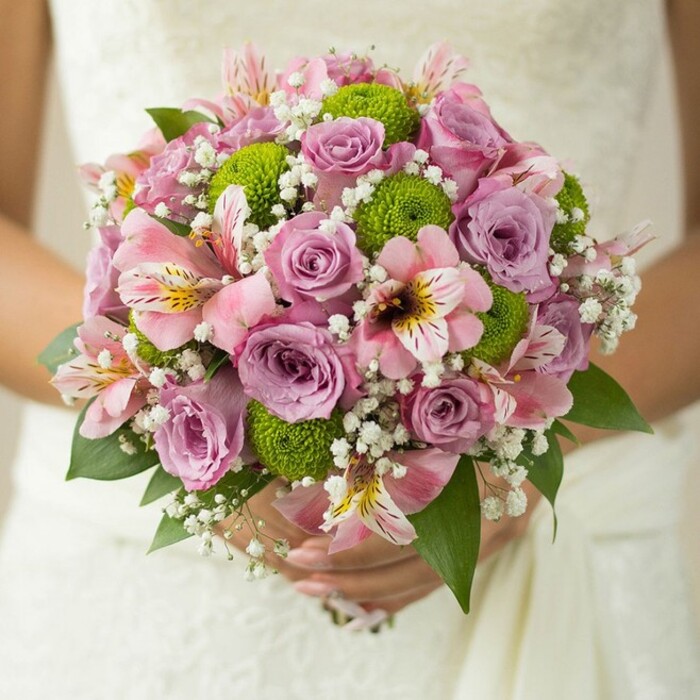 A radiant round bridal bouquet, lovingly held against the intricate lace of a wedding gown, reveals the thoughtful craftsmanship characteristic of Sands End florists. Central to the arrangement are soft lavender and pastel pink roses, their velvety petals opening in layers that catch the morning light, evoking both romance and celebration. Interspersed throughout are brilliant lime-green button chrysanthemums, their spherical forms lending a modern flare and burst of colour reminiscent of gardens along Townmead Road. Pink alstroemeria lilies-trumpet-shaped and alive with maroon and golden-striped markings-add a touch of exotic delicacy, while airy sprays of baby's breath thread throughout, imparting a sense of volume and lightness much like the open canal paths nearby. A few deep green, waxy rose leaves peek from the bouquet's base, providing contrast and structure. The hands of the bride, adorned with a simple wedding ring, cradle the bouquet as it rests against the soft fabric of her gown, creating an image of intimacy and anticipation. Crafted with local expertise by Florist Sands End, every element of this bouquet-from fresh, hand-picked blooms to the thoughtful interplay of colour and texture-reflects the joy and individuality of a wedding in this riverside neighbourhood. The overall impression is one of timeless elegance balanced with a cheerful hint of modernity, making it an exemplary accessory for spring or summer nuptials.