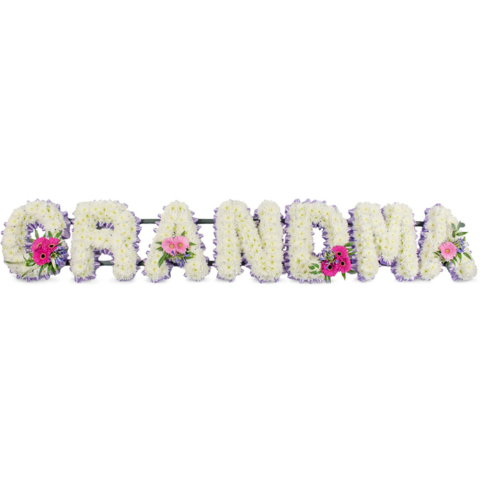 This photograph captures a long, horizontal funeral tribute where the word "GRANDMA" is spelled out in large capital letters, each letter deeply cushioned with pristine white chrysanthemums. The blooms are tightly packed and evenly sized, creating a smooth, velvety surface that looks almost cloudlike, inviting thoughts of softness and gentle touch. Each letter is neatly traced with a band of pale lilac ribbon, the satin edge catching the light and giving the outline a subtle shimmer that frames the pure white centres. The tribute rests against a neutral backdrop, so all attention is drawn to the flowers themselves - their rounded petals, the tiny yellow-green eyes of some of the chrysanthemums just peeking through, and the thoughtful placement of accent flowers. Three small posies sit along the word: at the opening "G", one of the central "A"s, and at the final "A". In each, bright pink gerbera daisies stand forward like little suns, accompanied by soft pink roses and miniature rosebuds, all woven with sprigs of fresh greenery that add natural depth and movement. The foliage, in varied shades of green, softens the white expanse and offers a quiet sense of life continuing. Imagined in the context of a Sands End farewell, perhaps after a family gathering near South Park or along Townmead Road, this tribute feels like a gentle final word spoken in flowers. It expresses gratitude, love, and the lasting imprint of a grandmother's presence, rendered through calm colour, careful craftsmanship, and a soothing, balanced design.