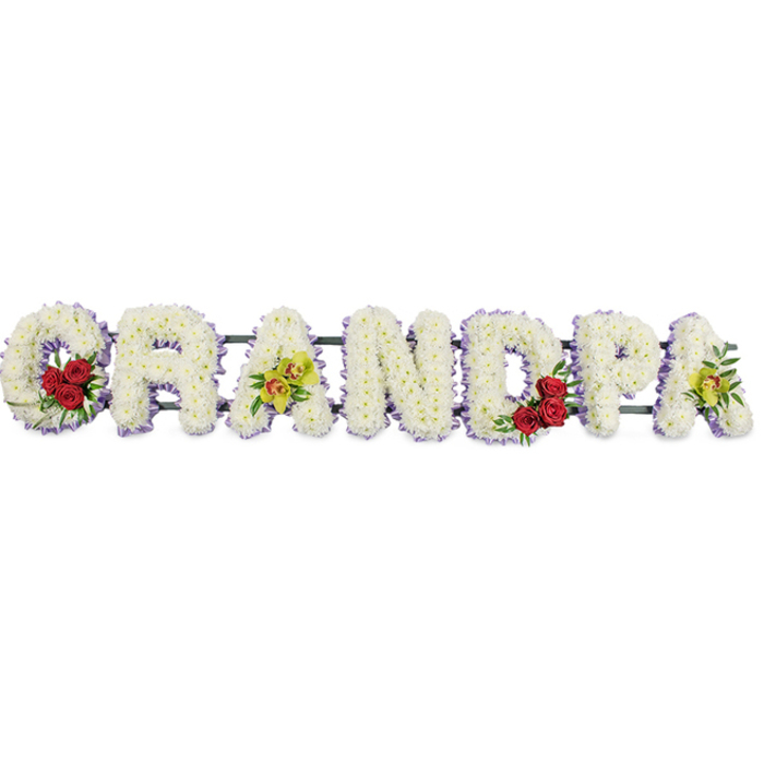 The image shows a horizontal funeral tribute that spells out the word "GRANDPA" in large, block-style floral letters, set cleanly against a plain white background so that every colour and contour can be clearly imagined. Each capital is carefully packed with pristine white chrysanthemums, their compact, cushion-like flower heads overlapping to create a plush, uniform surface that feels both soft and reverent, like a gentle blanket of petals laid in honour of a cherished grandfather. Around the outer edge of each letter, a slim band of pale lavender ribbon offers a soft hint of colour, outlining the shapes and lending a quiet elegance reminiscent of calm evening skies over nearby Sands End Park. At the beginning of the word, cradled inside the curl of the "G," sits a small but striking cluster of deep red roses, framed with glossy green foliage, providing a strong focal point that speaks of deep affection and respect. The third letter, the first "A," carries a spray of yellow-green orchids, their delicate, exotic blooms introducing a lighter, modern touch that many local families around Sands End and Imperial Wharf choose for personalised tributes. In the curved inner space of the "D," another grouping of three or four red roses mirrors the "G," creating a balanced rhythm of colour, while the final "A" again features the same soft yellow-green orchids, tying the design together visually. Slim, dark support bars quietly hold the letters in a straight line, ready to be placed beside a coffin or at the front of a service. The overall effect is one of calm purity, gentle colour accents, and sincere remembrance, perfectly suited to saying goodbye to a beloved grandfather with grace and dignity.