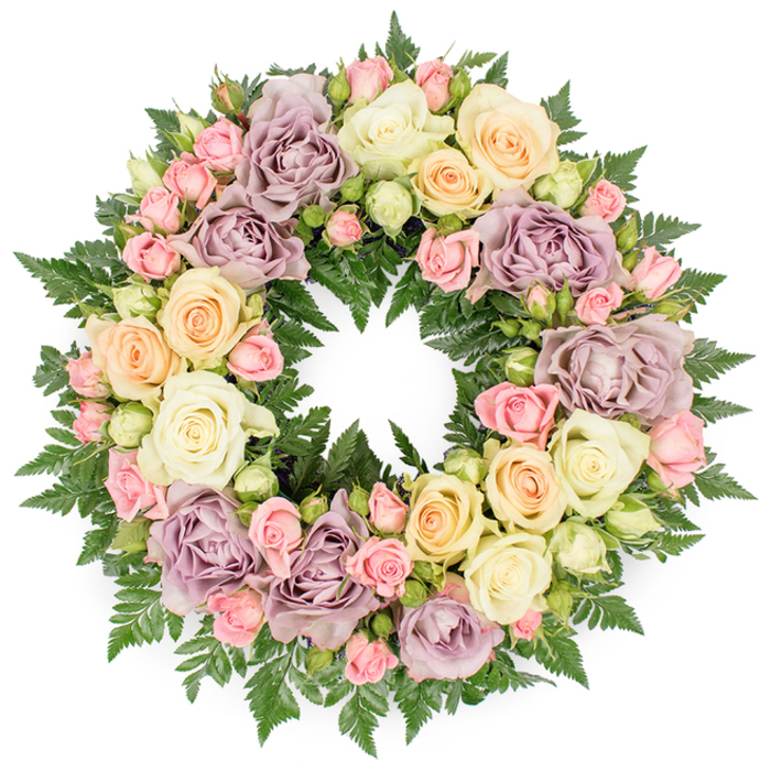 A classic circular sympathy wreath is displayed in full view against a crisp white background, its form completely defined by roses in pastel hues and a halo of rich green foliage. The wreath is dense and opulent, made up of many layers of blooms that overlap to create a soft, quilted surface of petals. Large roses in shades of creamy white and gentle ivory dominate the design, their fully opened centres revealing intricate spirals that feel calm and reassuring. Scattered between these are delicate clusters of small pink spray roses, some tightly closed, others just beginning to open, bringing a sense of tenderness and unfolding memory. Subtle threads of pale peach and buttery yellow roses introduce a warm, comforting light, while distinctive dusty lavender and muted mauve roses lend an elegant, antique note that suggests reflection and quiet dignity. Beneath and between the roses, finely textured dark green fern fronds and similar foliage form a lush base, their feathery tips protruding just enough to frame the flowers and add depth, contrast, and a sense of natural movement. The circle is perfectly formed, with the central opening clearly visible, reinforcing the symbolism of eternity, continuity, and the unbroken bond between the living and the departed. Even under soft, neutral lighting, the pastel roses appear fresh and dewy, as if newly arranged for a same-day farewell service. This type of wreath could be lovingly created in a Sands End florist's studio for a funeral, memorial, or graveside tribute, offering families a peaceful, beautifully crafted expression of remembrance and sincere condolences.