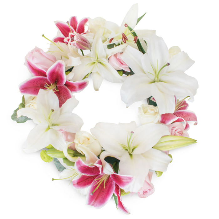 Against a crisp, white backdrop, this elegantly circular floral wreath captures the heartfelt spirit of remembrance cherished in the Sands End community. Dominating the design are luxuriant Oriental lilies, their star-shaped pure white blooms unfurled gracefully to reveal luminous green stamens and subtle golden pollen. Well-placed among these are spectacular fuchsia-pink stargazer lilies, each petal alive with a vibrant magenta hue, delicately flecked with crimson spots and outlined in white-their lively contrast bringing an almost celebratory vibrance to the arrangement. Clusters of romantic blush pink and cream roses, ranging from tightly closed buds to open, ruffled flowers, rest between lilies, imparting a tender, textural softness. Gentle wisps of eucalyptus and polished green foliage weave through the circle, imbuing the wreath with a fresh, natural structure that echoes the tranquillity of nearby riverside walks. The overall effect is one of luminous abundance-a seamless, unbroken ring symbolic of eternal affection and reverence. Every flower and leaf is thoughtfully placed, displaying the craftsmanship and care familiar to those who order from local Sands End florists for services at St. John's Church or commemorations near Hurlingham Park. The design's serene colour palette, tactile variety, and lush fullness create a fitting tribute for sympathy, funerals, or celebration of life ceremonies, allowing families to express grief, love, and remembrance with grace.