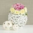 A delicately styled floral composition rests on a smooth white surface, as if awaiting guests at an intimate wedding reception in Sands End. The centerpiece focuses on a rounded bouquet of roses, about a dozen in total, placed tightly together to form a plump, dome-like shape with no stems in sight. The upper blooms glow in tones of pastel pink, swinging from soft blush to gentle lavender-fuchsia, while the lower layer carries creamy butter-yellow and ivory hues, so that the colours gently band together without sharp divisions. Each rose appears freshly cut, petals unblemished and velvety, some tightly swirled in bud-like form, others gently unfolding to reveal their layered hearts. Fine, airy sprigs of white baby's breath lace between the larger flowers, their many tiny blossoms catching the light and bringing a sense of lightness and movement to the design. This floral dome is set in a distinctive, spherical vase made from slender, white-painted branches or wicker, woven into an open, basket-like structure. The latticework reveals glimpses of the inner container and casts delicate shadows across the tabletop, adding a tactile, handcrafted quality. To the left, two decorative balls fashioned from pale natural wood echo the open-weave pattern in a more rustic tone, one medium-sized and one small, arranged casually yet harmoniously. Behind the arrangement, a softly textured beige or light taupe background, reminiscent of linen or subtle wallpaper, recedes quietly, making the pastel roses and crisp white elements stand out. The overall impression is one of gentle romance and modern simplicity, well suited to a wedding table arrangement, bridal brunch, or vow renewal celebration styled by a Sands End florist who prizes graceful details and calm, welcoming beauty.