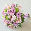 A lush, rounded bouquet bursts alive with pastel pinks, lilacs, and vivid greens, thoughtfully composed by a Sands End florist and presented against a soft, neutral background. At the heart of this arrangement, roses bloom in gentle shades of mauve and blush, their petals unfolding in elegant spirals that convey charm and sophistication-much like the hidden gardens that dot Sands End. Interspersed are vibrant lime green clusters of button mums, adding contemporary vibrance and textural playfulness to the pastel canvas. Pale pink alstroemeria lilies, with petals accented in yellow and streaked with deep maroon, bring graceful movement and depth, while frothy white sprigs of baby's breath (gypsophila) weave a delicate, ethereal mist throughout the bouquet. Rich green leaves peek between the blooms, enhancing the composition's fullness and vitality. The tightly bound stems are wrapped in a lustrous pink satin ribbon, a hint of celebration glimmering in the soft light, finished with a delicate strand of pearls for a touch of bridal romance. The bouquet's harmonious blend of colours and textures makes it an ideal centerpiece for weddings along the Thames or gifts for cherished occasions within Sands End, celebrating love, elegance, and the personal artistry of local floristry.